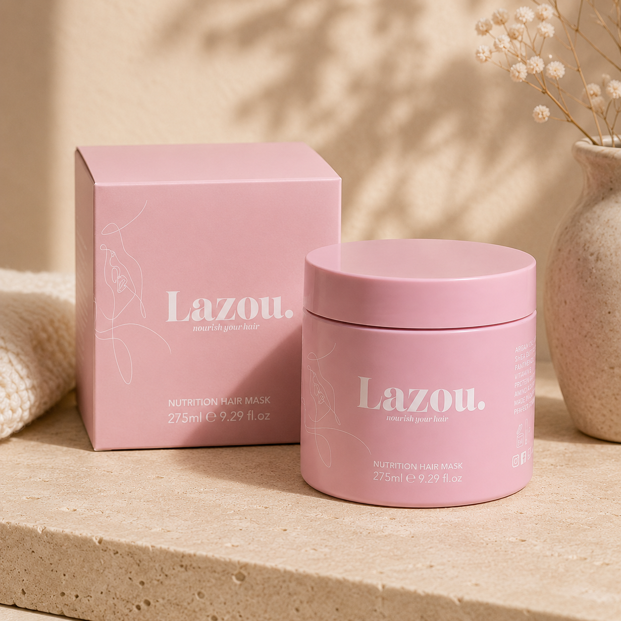 A pink jar of Lazou. Nutrition Hair Mask stands next to its matching pink box on a beige stone surface, with soft shadows and dried flowers in the background.